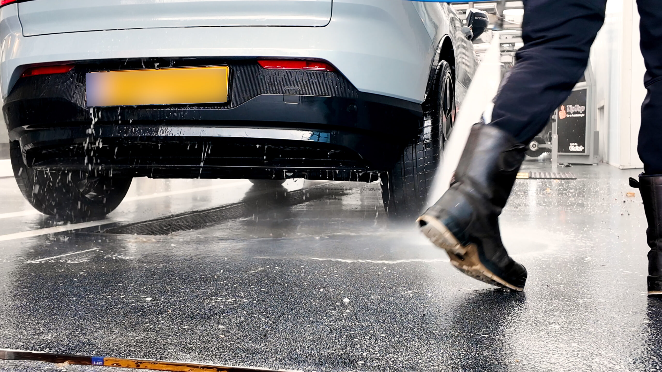 Afspoelen van de onderkant tijdens het wassen Achterkant van een auto die wordt afgespoten met water tijdens het wasproces, met een medewerker in werklaarzen op de vloer