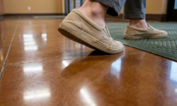 Persoon loopt over glimmende, gladde vloer Voeten in beige slip on schoenen lopen over een glanzende bruine vloer, met een groene deurmat op de achtergrond