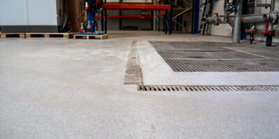Praktijkbeeld van een industriële vloer met afvoergoten Grijze gespikkelde industrievloer met metalen afvoergoten en rooster, opslagstellingen en leidingwerk op de achtergrond