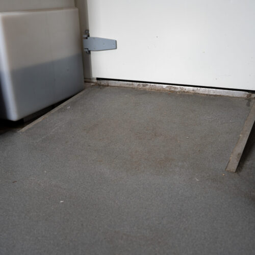 Grijze antislip vloer met lichte verzakking bij drempel van koelceldeur, met een krat op wielen ernaast