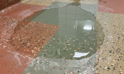 Blijvende plas water door slechte afwatering in sanitaire ruimte Gespikkelde terrazzovloer in rood, oker en groen in sanitaire ruimte met een grote plas water die niet wegloopt
